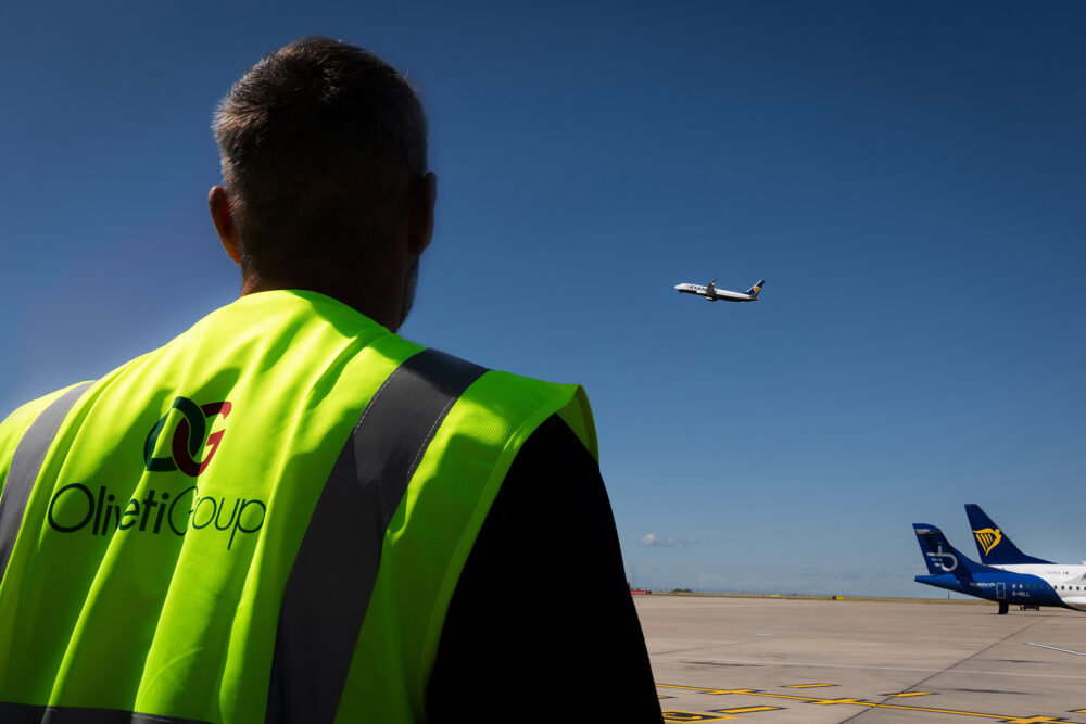 A man in Oliveti high-vis looking at a plane taking off.